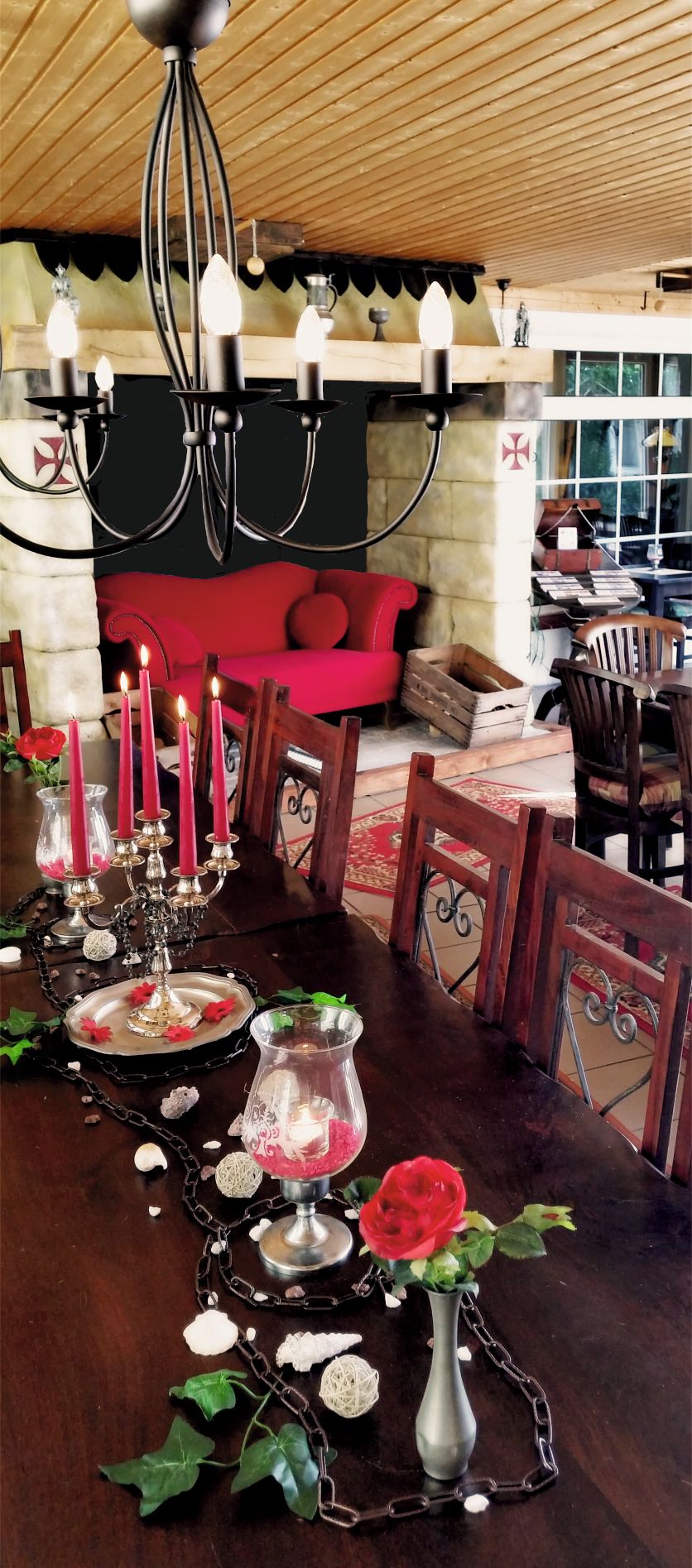 Un comedor con una mesa de madera dispuesta para una comida, con una araña, velas y un florero con una rosa roja.