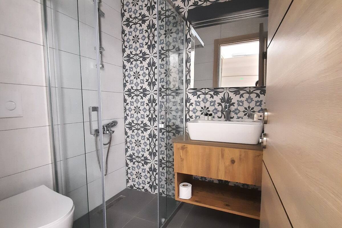 Un baño moderno con cabina de ducha de vidrio, inodoro blanco, tocador de madera con lavamanos y azulejos con diseño.