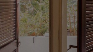 Una habitación con ventana, persianas de madera y vistas a árboles y una valla.