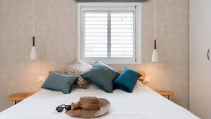 Un dormitorio con una cama, almohadas, un sombrero y gafas de sol sobre la cama. Hay dos mesitas de noche y una ventana con persianas.