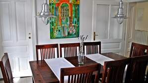 Een eethoek met een houten tafel en stoelen, een kleurrijk schilderij aan de muur, en twee hanglampen.