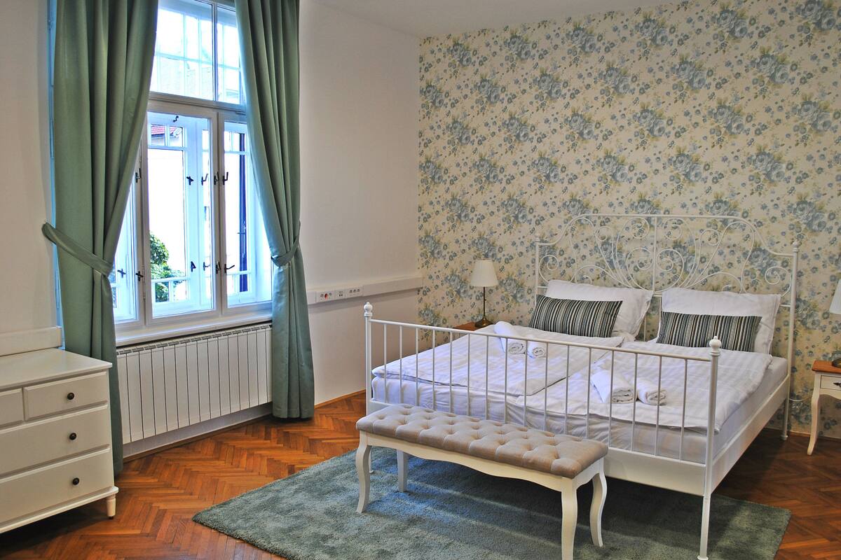 Une chambre à coucher avec un lit en métal blanc, un banc, une commode et une fenêtre avec des rideaux.
