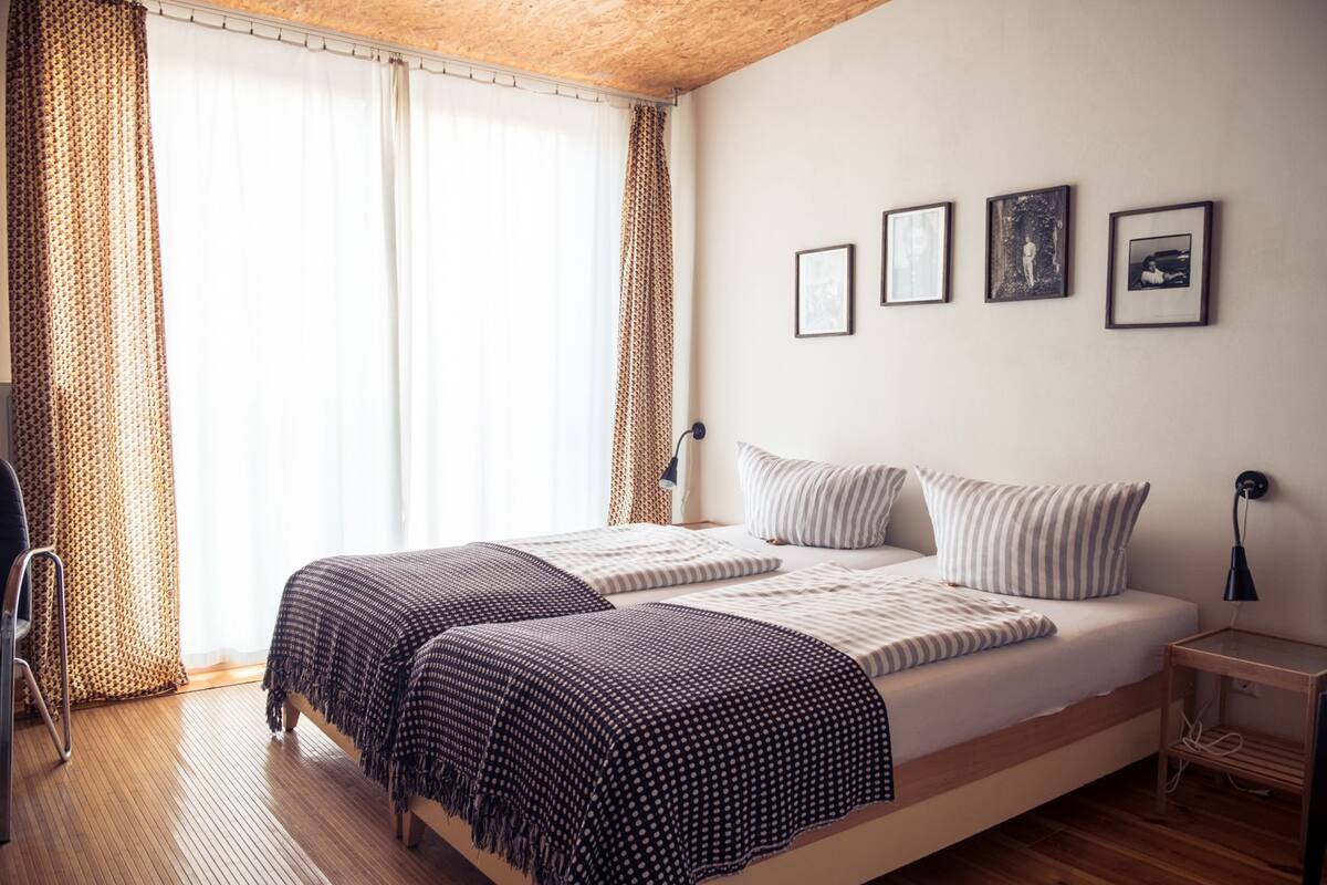 A double bed with striped bedding, a wooden floor, and a window with patterned curtains.