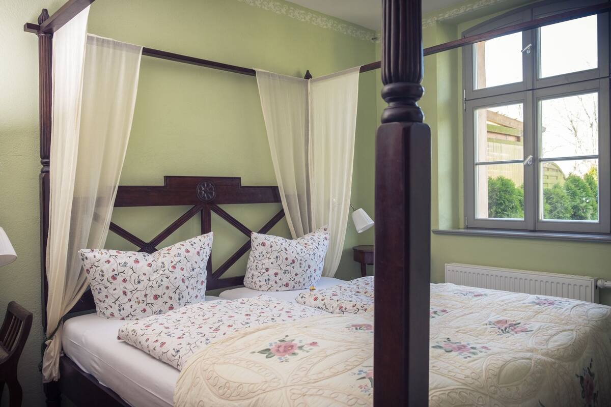A four-poster canopy bed with floral bedding and a view of greenery outside.