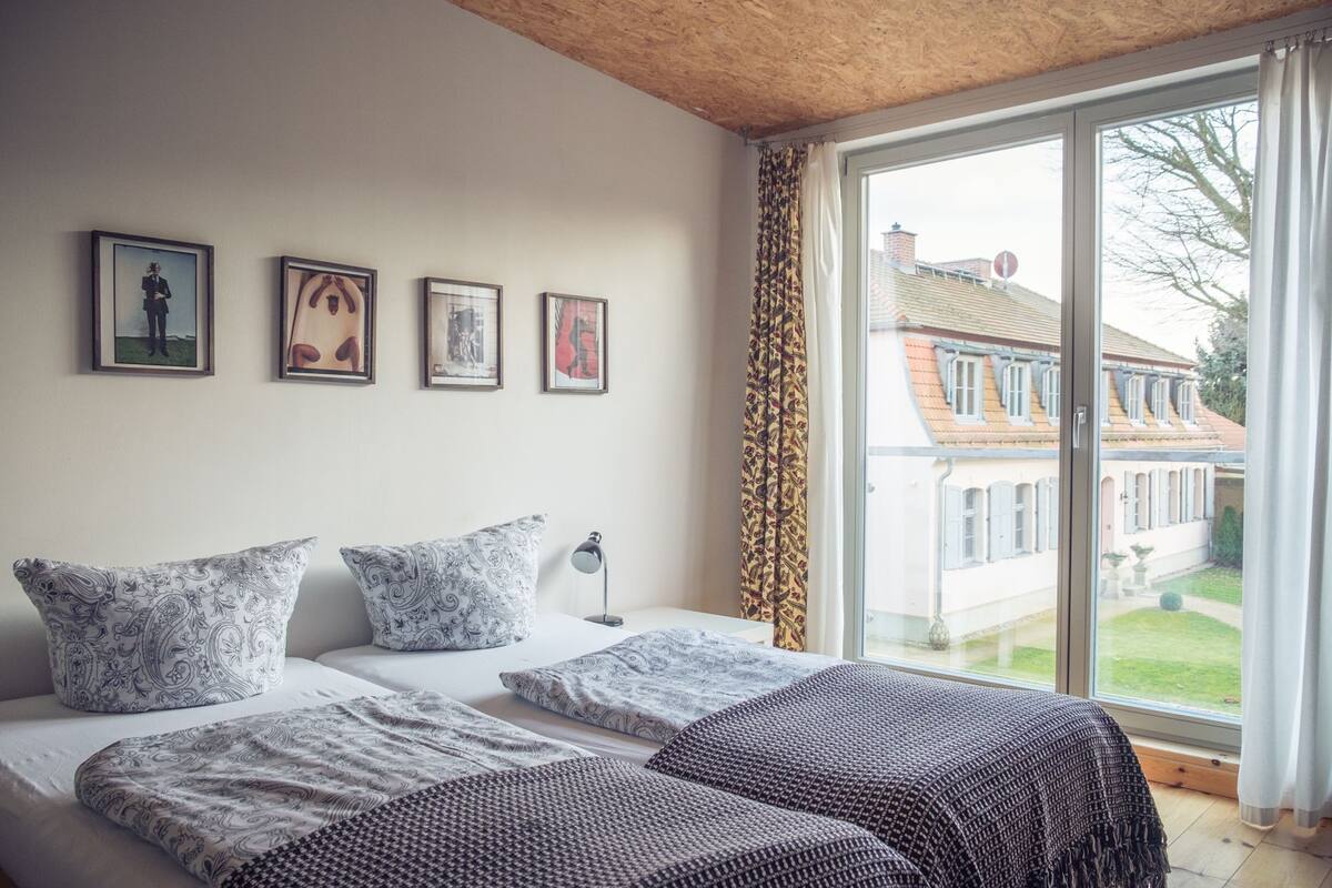 A bedroom with a large window, a bed with patterned bedding, and four framed pictures on the wall.