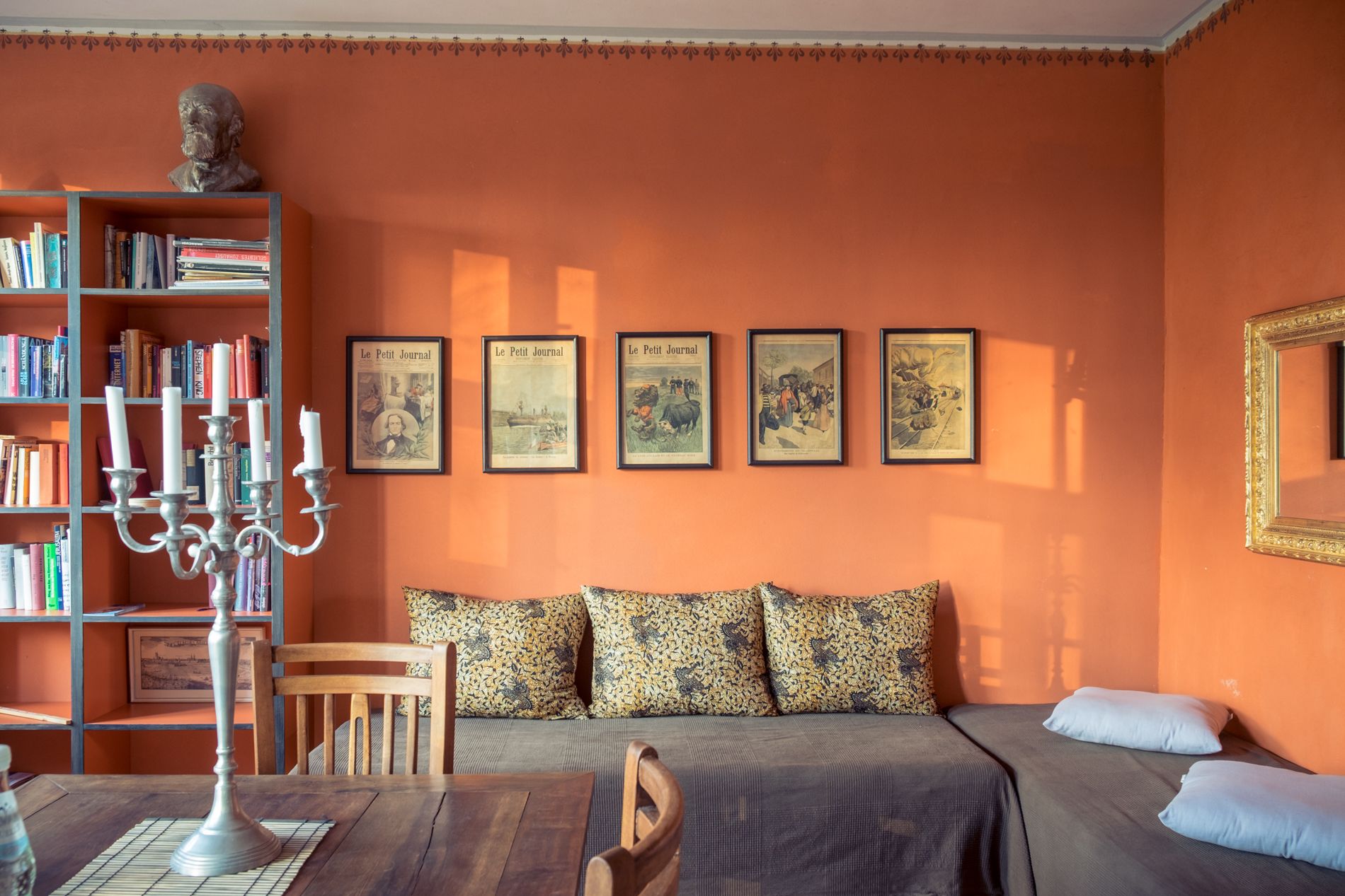 A cozy room with a bookshelf, a candelabra, a sofa with patterned cushions, and a bed with a blue pillow.