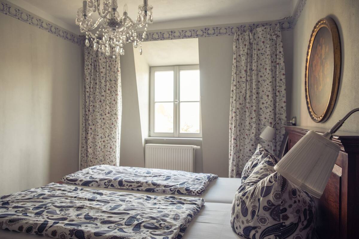 A bedroom with two beds, a chandelier, a window with curtains, a radiator, and a mirror on the wall.