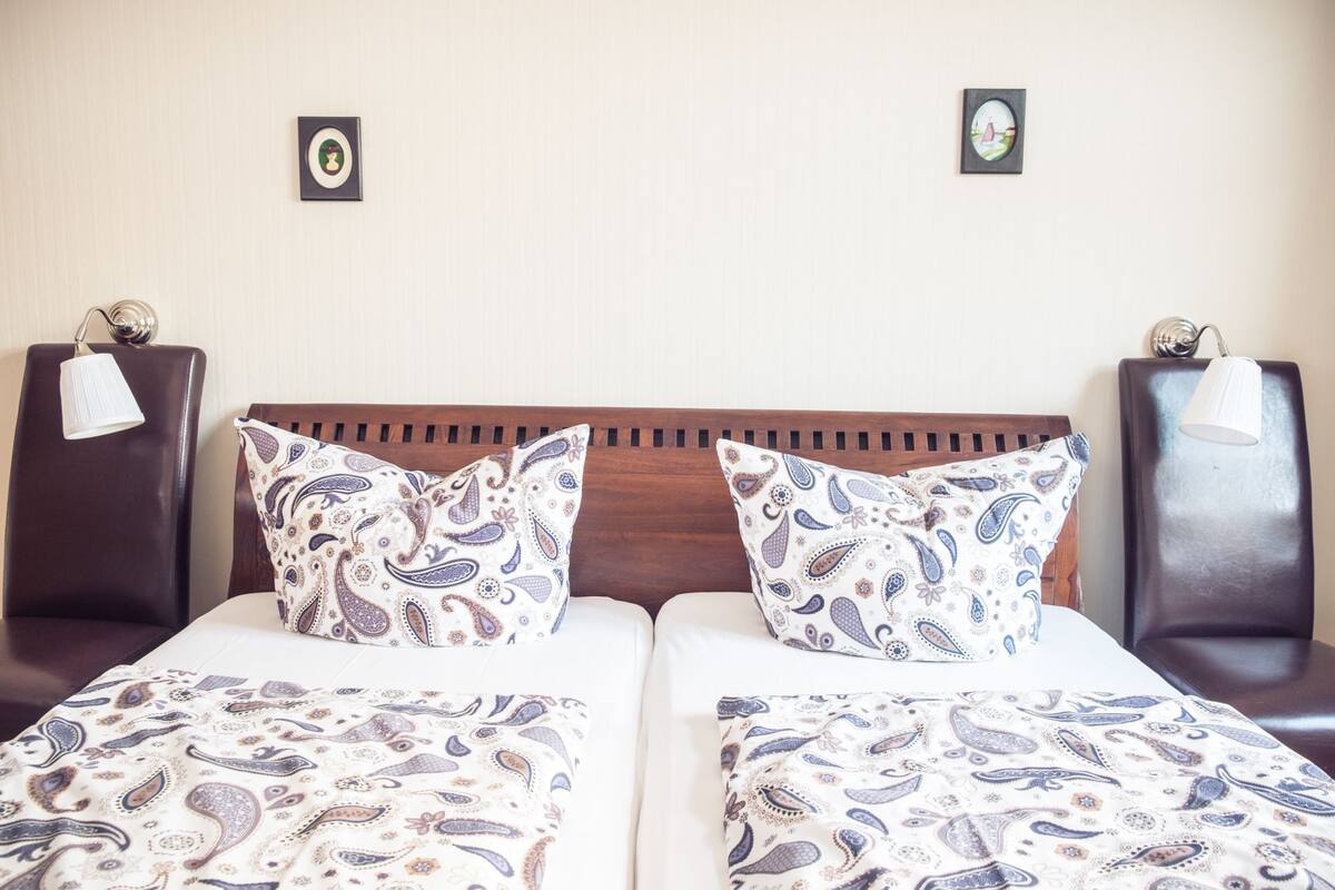 A hotel room with two beds, each with a patterned bedspread and matching pillows. There are two wall-mounted lamps on either side of the beds.