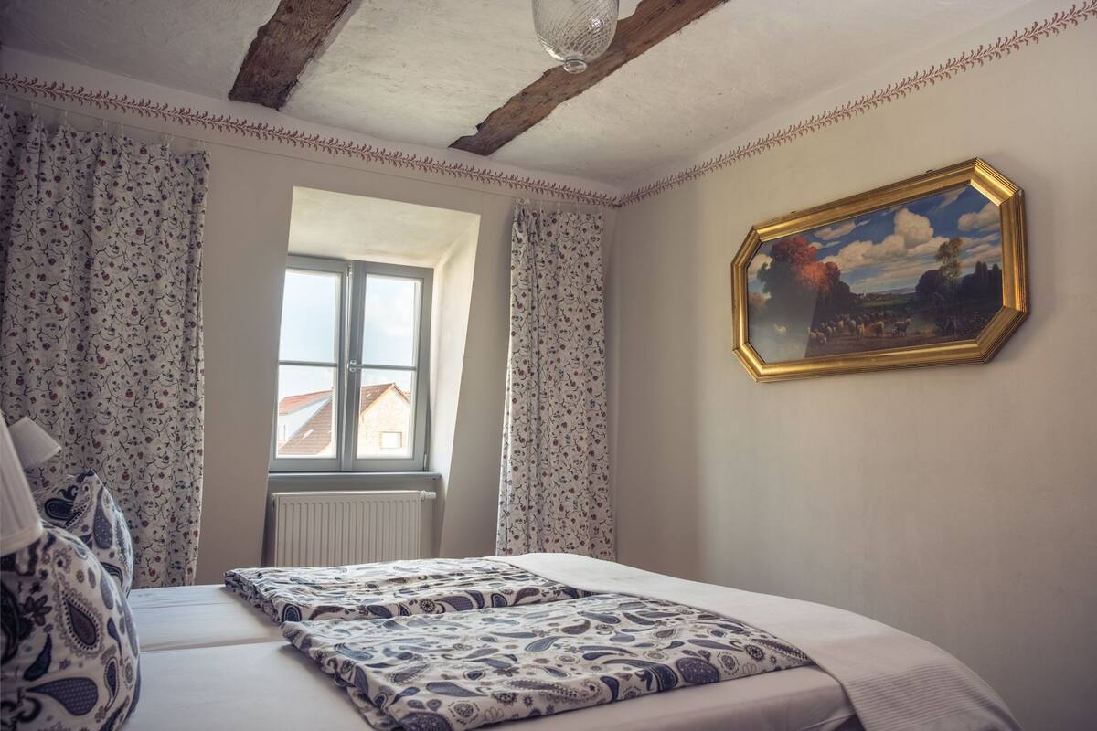 A bedroom with a large window, a bed with patterned bedding, a radiator, and a framed painting on the wall.