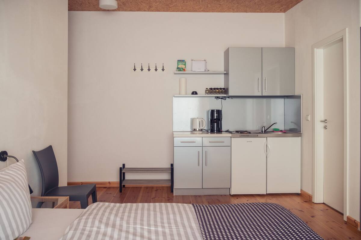 A compact bedroom with a bed, bedside table, and a small seating area. Adjacent is a kitchenette with a coffee maker, kettle, and cabinets.