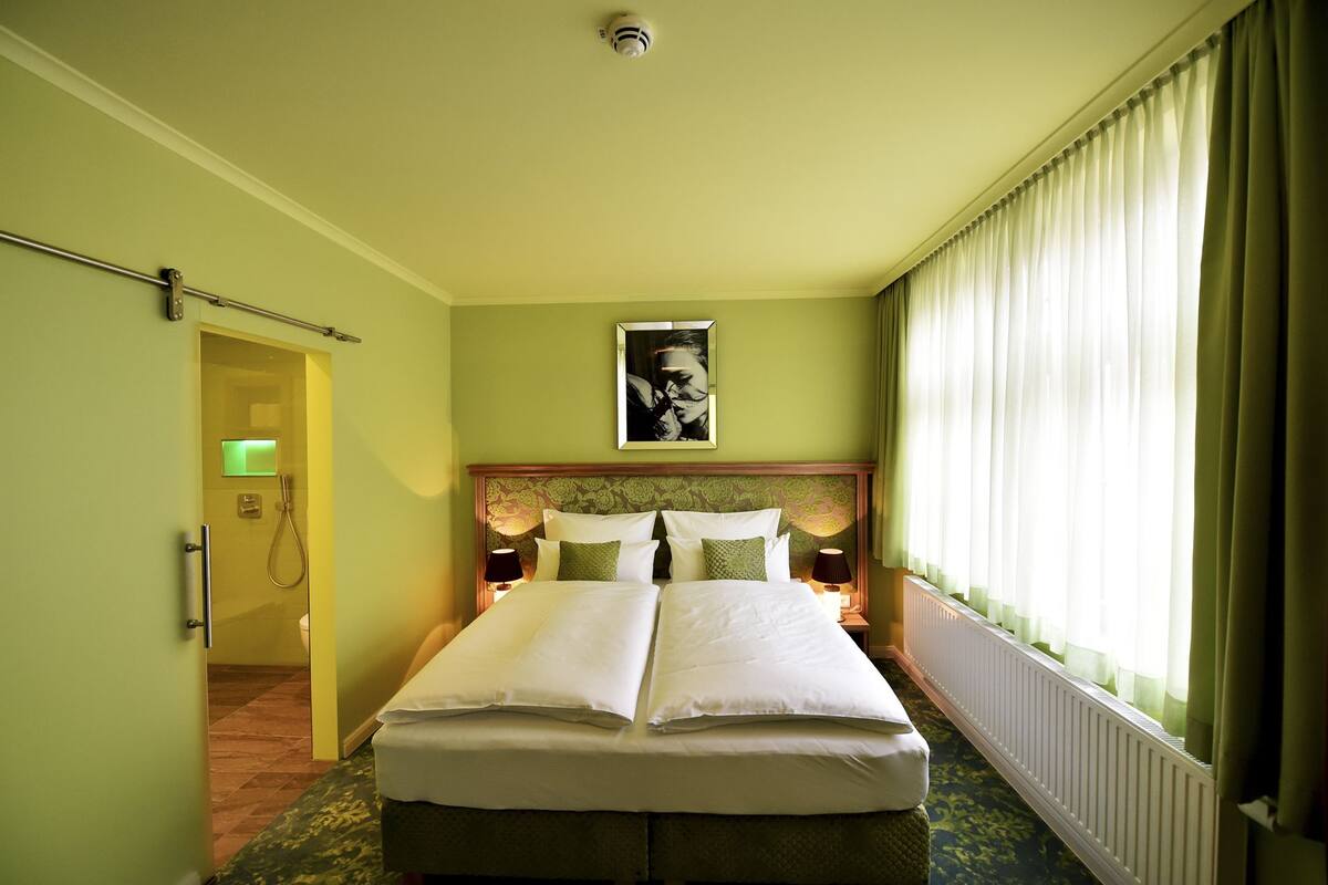 A hotel room with a double bed, a headboard with a floral pattern, a framed picture above the bed, a bedside table with a lamp, and a window with sheer curtains.