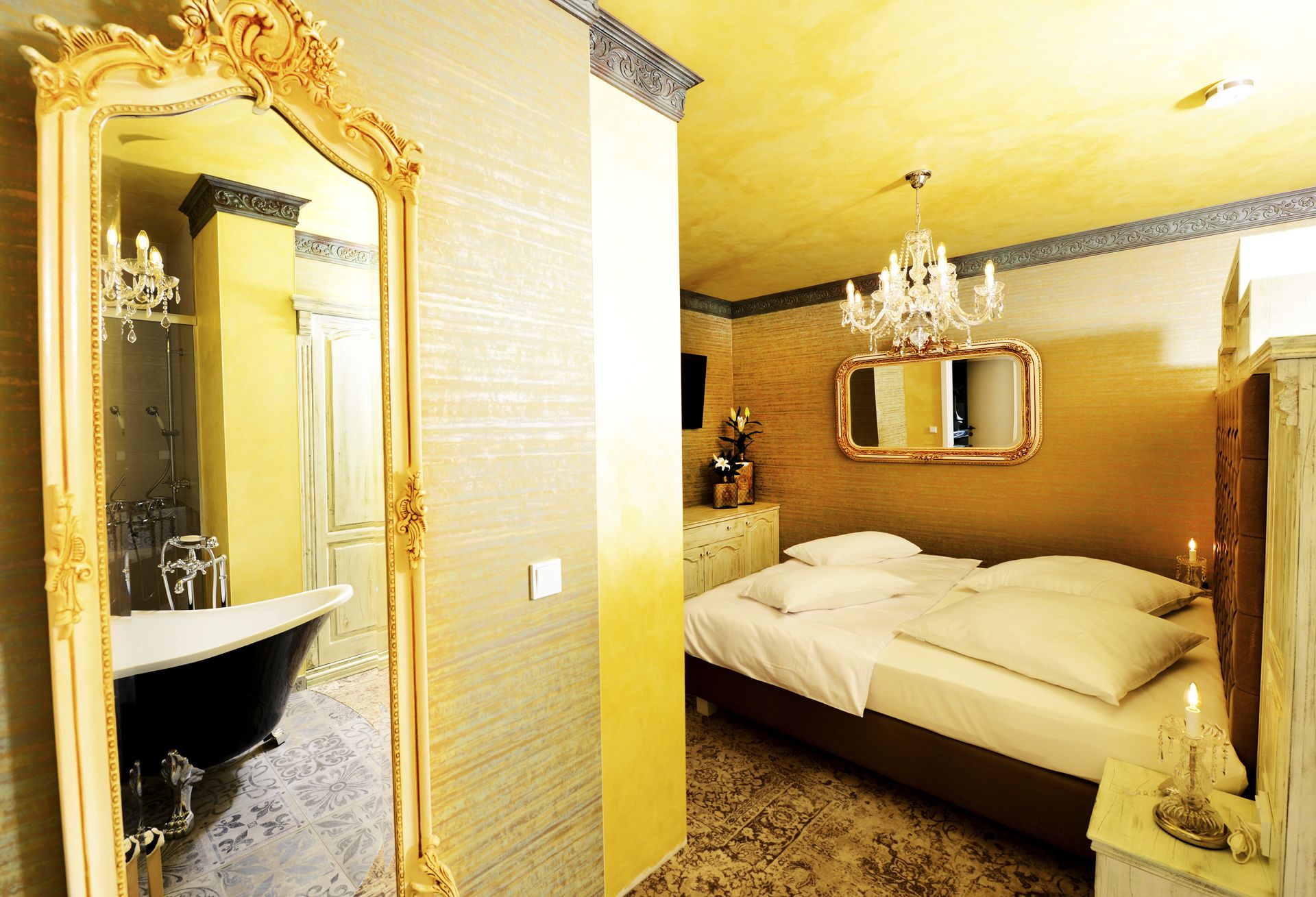 A room with a large ornate mirror, a bathtub, a bed with white linens, and a chandelier.