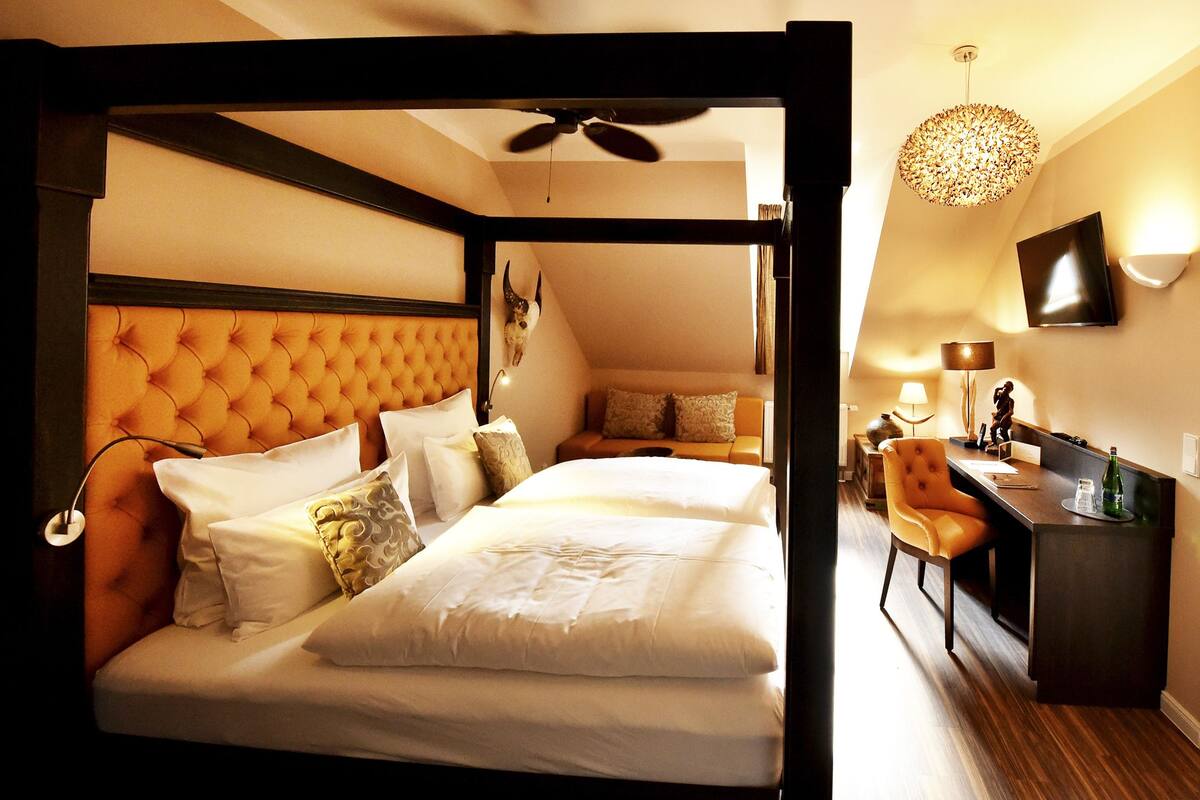 A hotel room with a tufted headboard, a bed with multiple pillows, a desk with a chair, a television, and a ceiling fan.