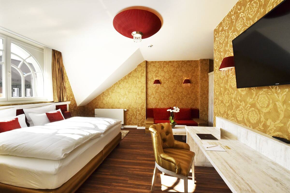 A hotel room with a large bed, a desk with a chair, a TV, and a red ceiling light.