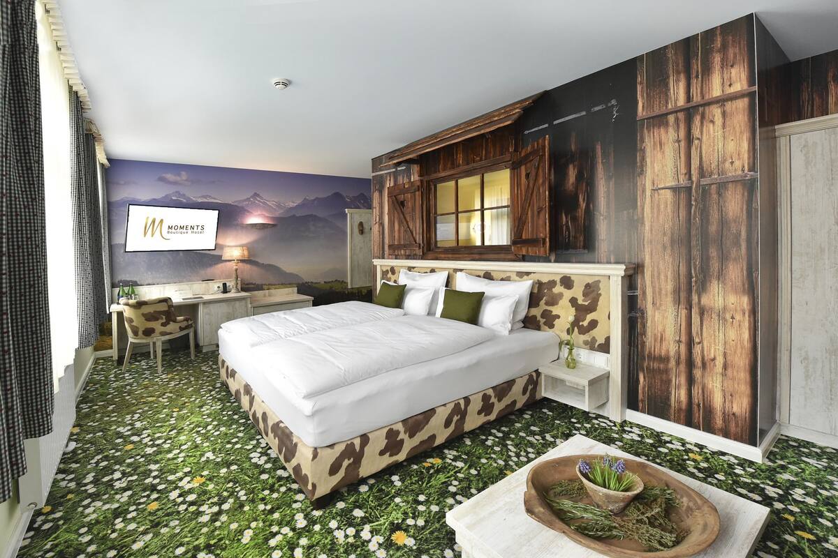 A hotel room with a bed, a desk, a chair, a wooden wardrobe, and a wall mural.