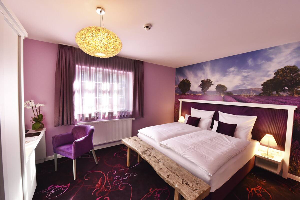 A bedroom with a bed, a chair, a desk, a lamp, and a painting on the wall.
