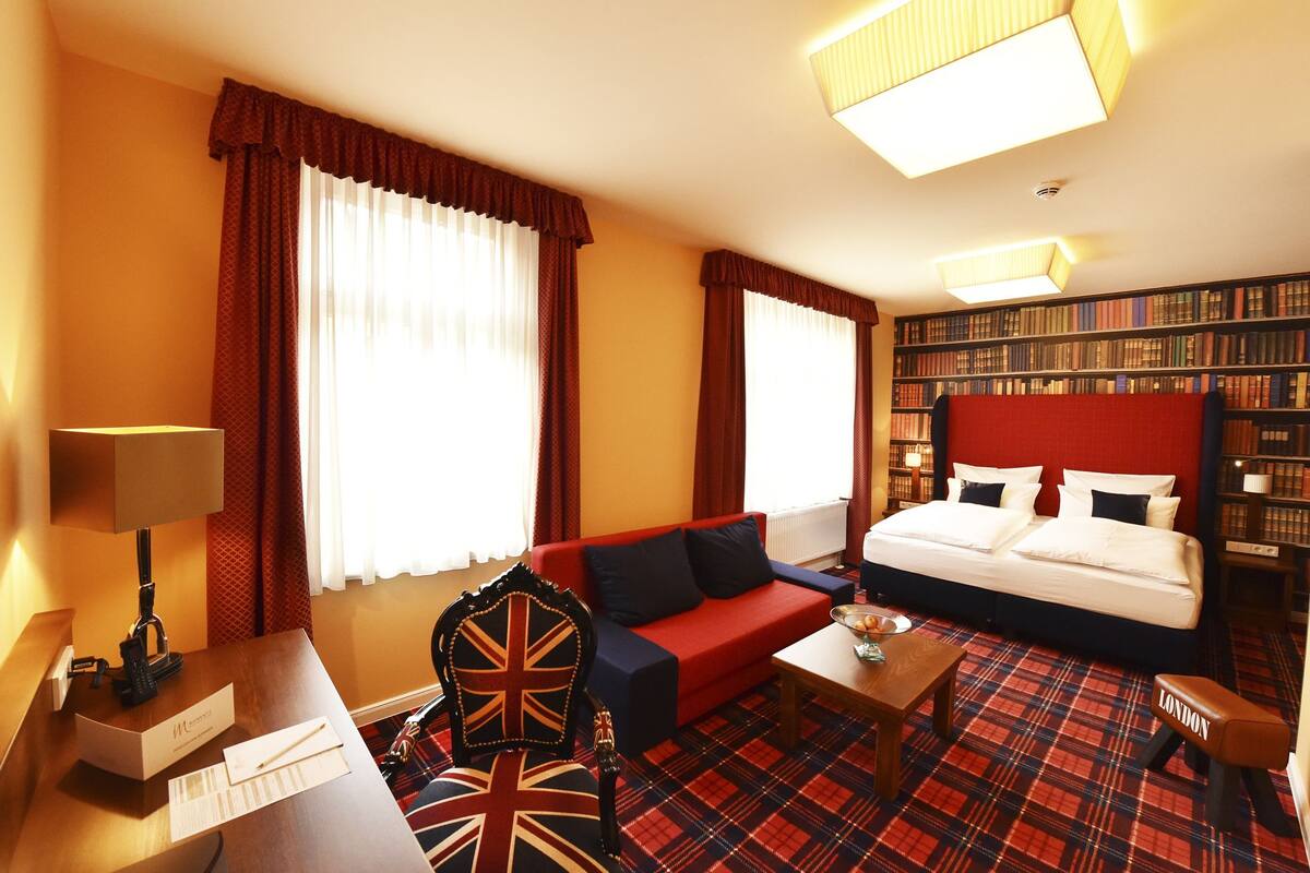 A hotel room with a bed, a red sofa, a coffee table, a chair with a Union Jack design, a bookshelf, and a red plaid carpet.