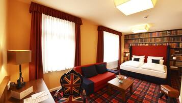 A hotel room with a bed, a red sofa, a coffee table, a chair with a Union Jack design, a bookshelf, and a red plaid carpet.