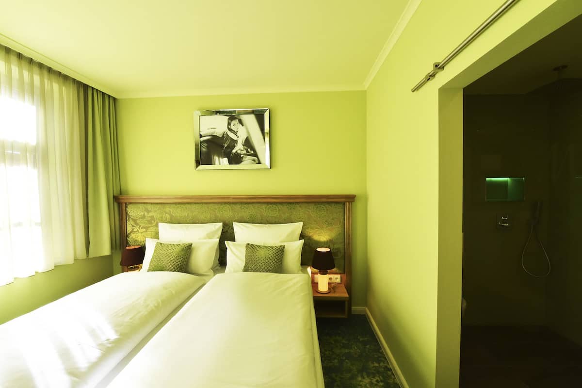 A hotel room with a bed, two bedside tables, a nightstand, a framed picture on the wall, and a shower area.