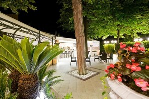 An outdoor patio area with a dining table and chairs, surrounded by potted plants and a large tree. - Hotel Belsoggiorno (Rimini)