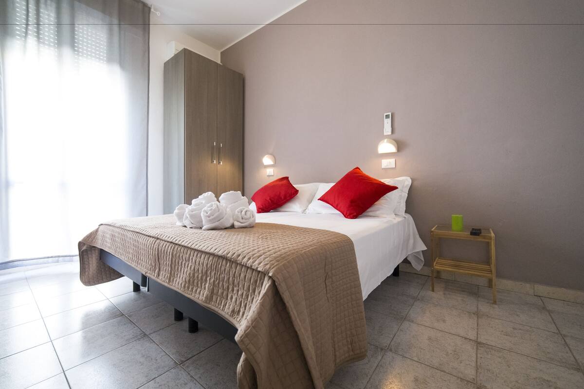 Una habitación de hotel con una cama, un armario de madera, una mesita de noche con una taza verde y un ventanal con cortinas de encaje.