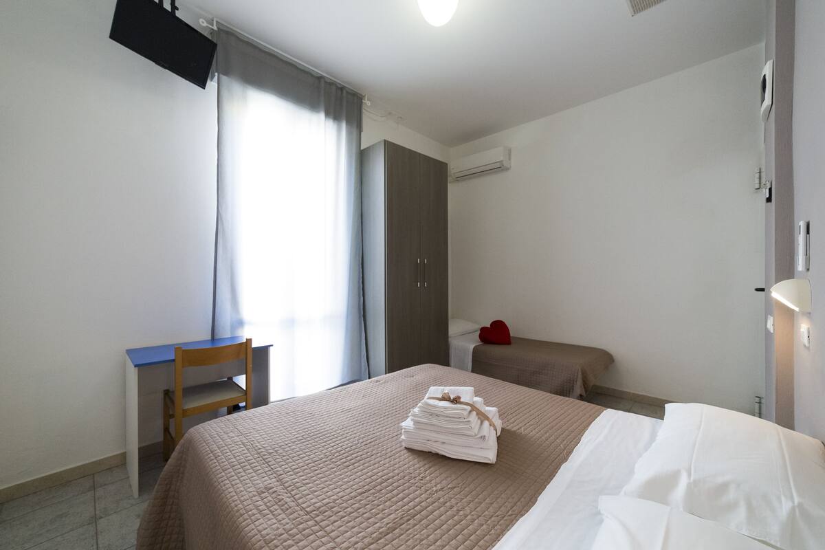 Habitación de hotel con cama, escritorio, silla, armario y mesita de noche con un objeto rojo.