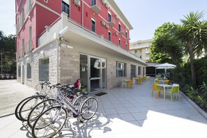 Un edificio con fachada roja, una zona de estar al aire libre cubierta con mesas y sillas amarillas y blancas, y bicicletas estacionadas afuera.
