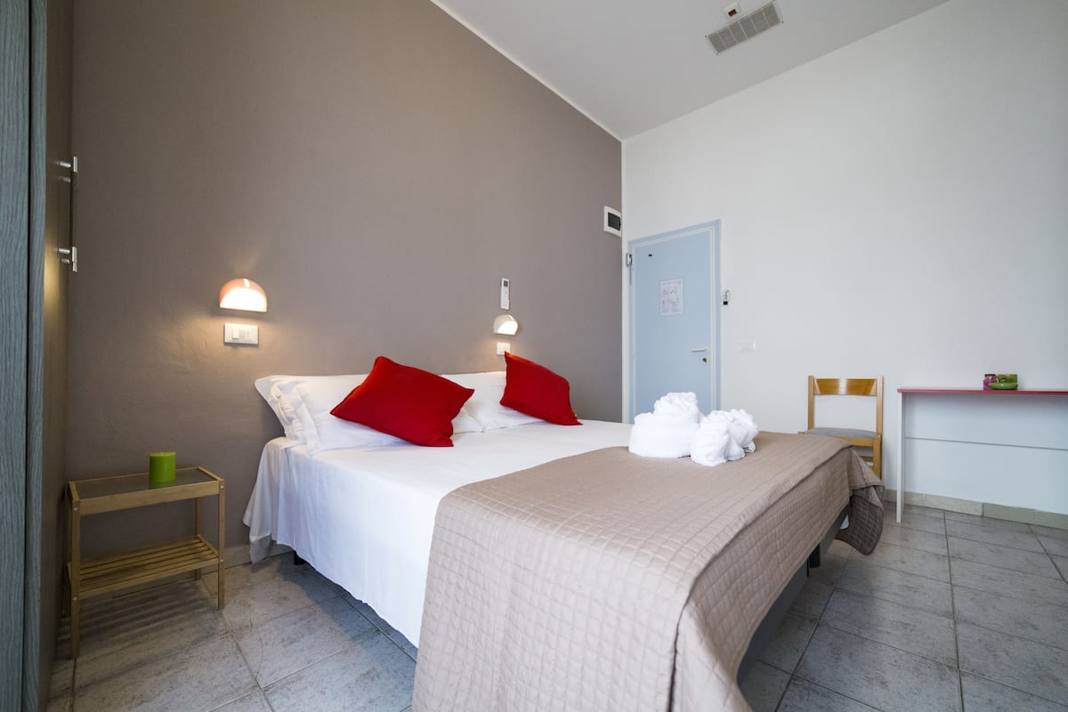 Un dormitorio con una cama, dos lámparas de noche, una mesita de noche de madera, una silla, un escritorio y una puerta.