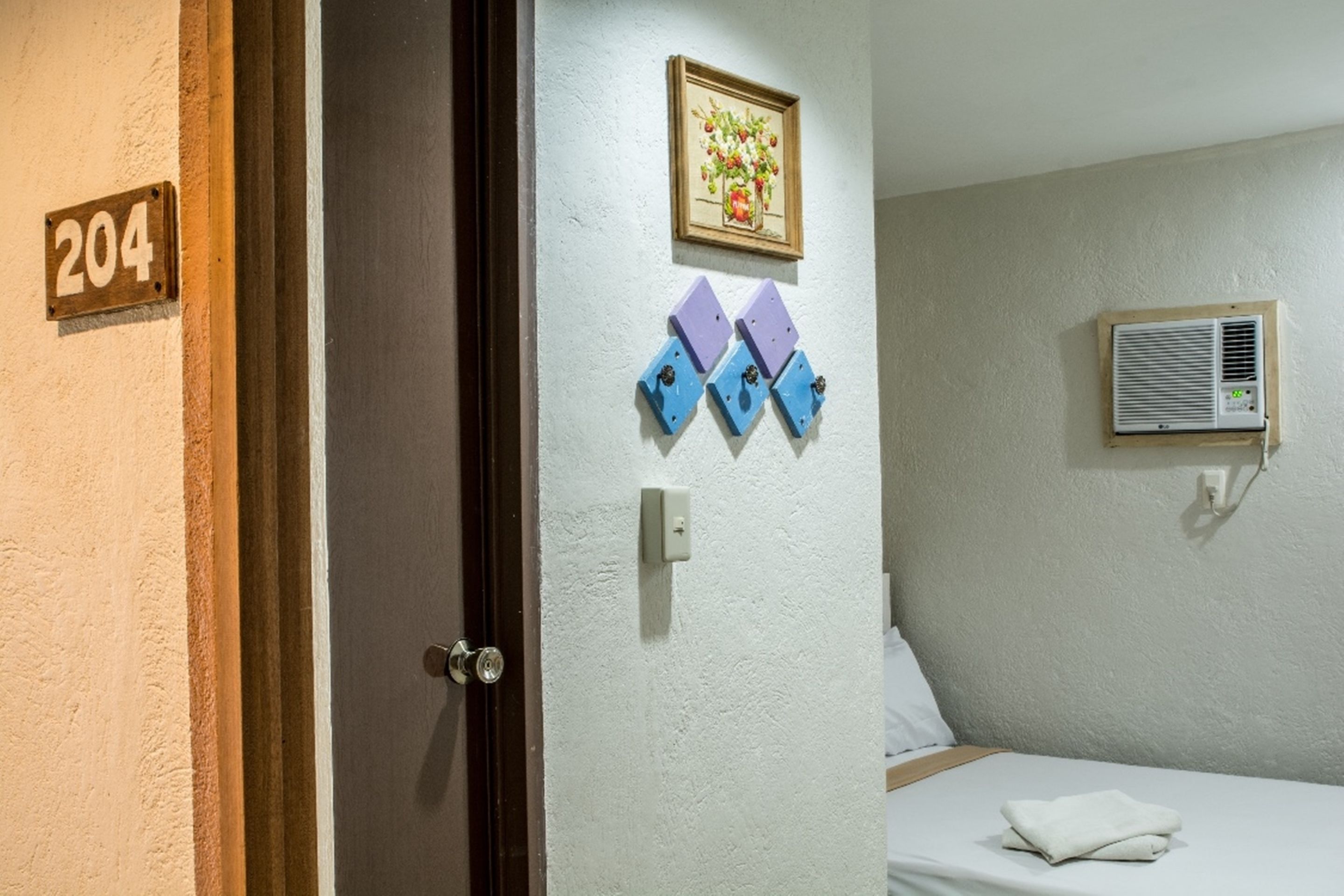 Habitación de hotel con una puerta que indica el número 204, una cama con almohada blanca, aire acondicionado montado en la pared y un cuadro enmarcado sobre la cama.