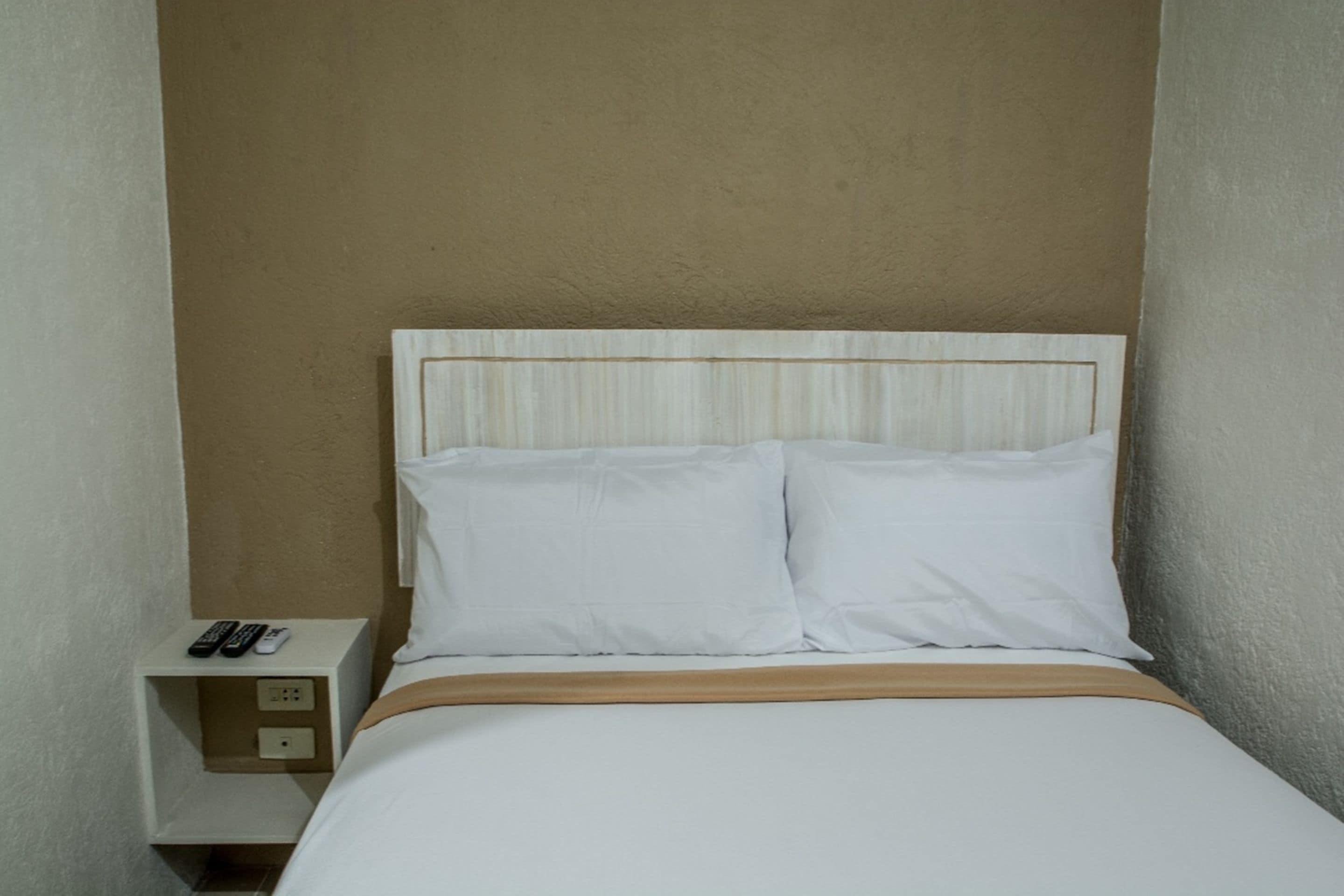Un lit simple avec une literie blanche et une tête de lit en bois. Il y a une table de chevet équipée d’une prise électrique et de deux télécommandes.