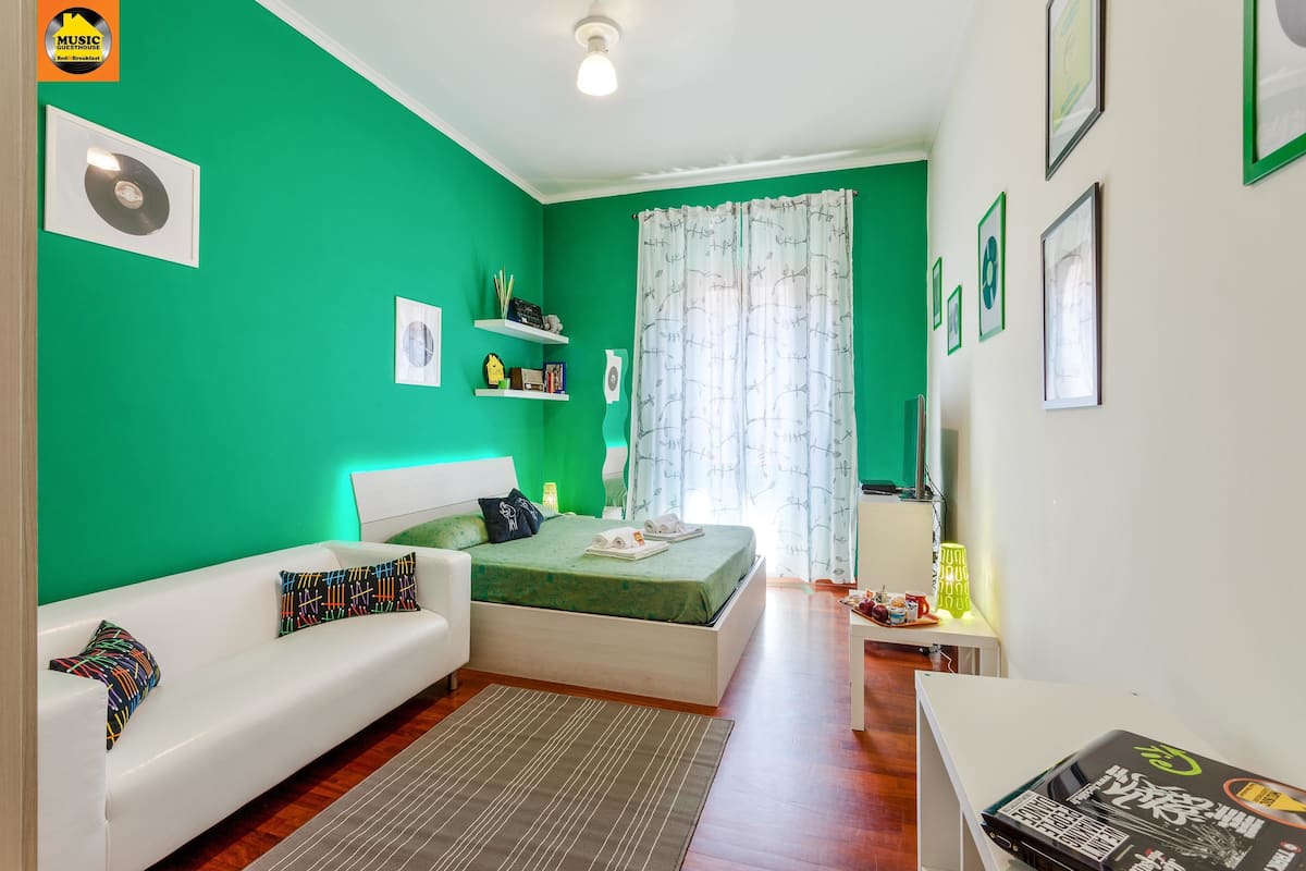A room with a green wall, a white sofa, a bed, and a small table with a lamp.