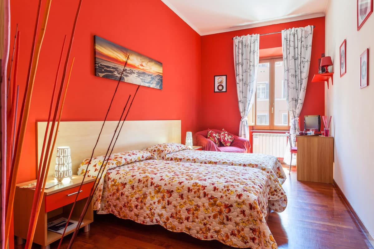 A bedroom with a red wall, a bed with floral bedding, a nightstand, a desk with a computer, and a window with curtains.