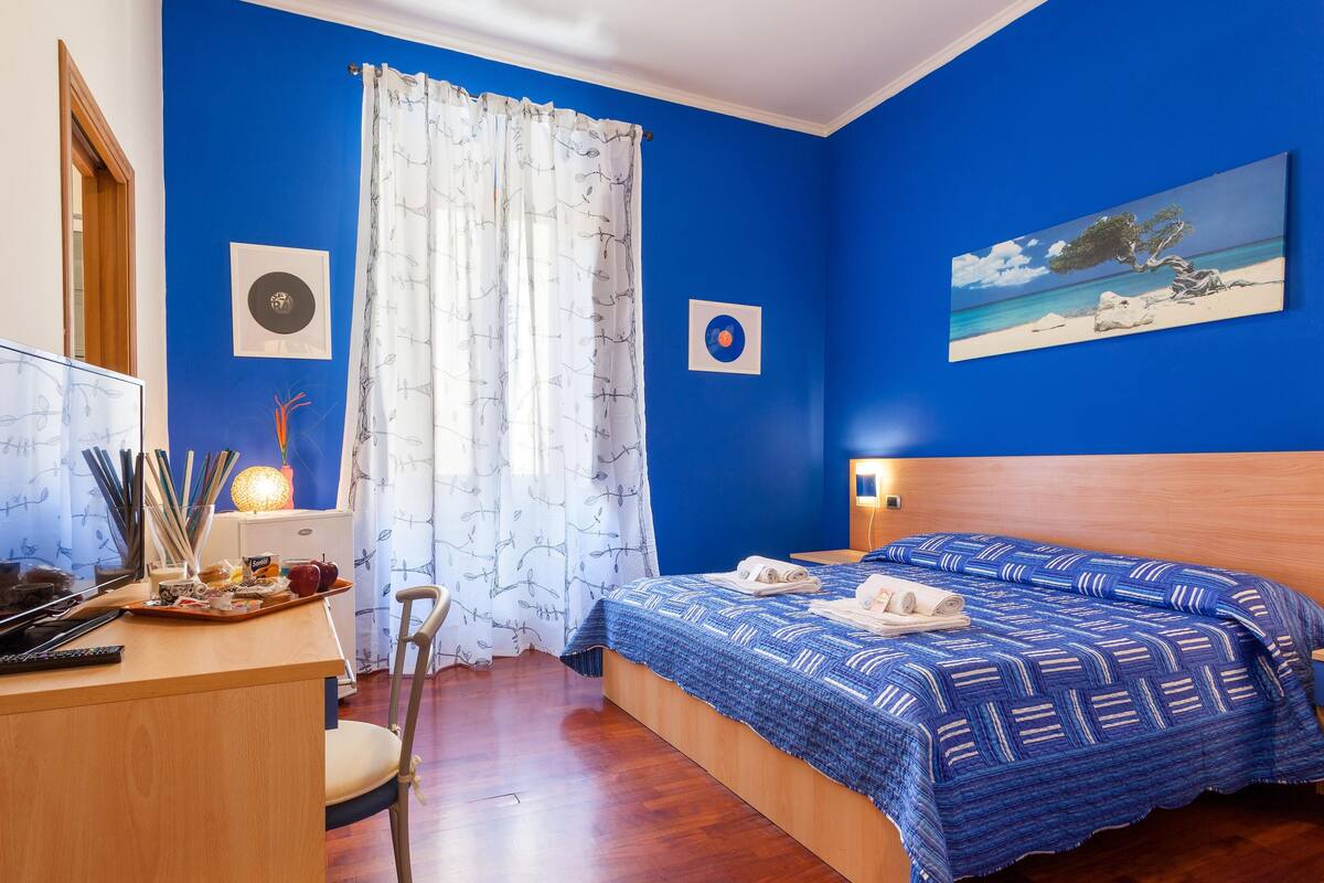 A room with a bed, a desk, a chair, a window with curtains, and a painting on the wall.