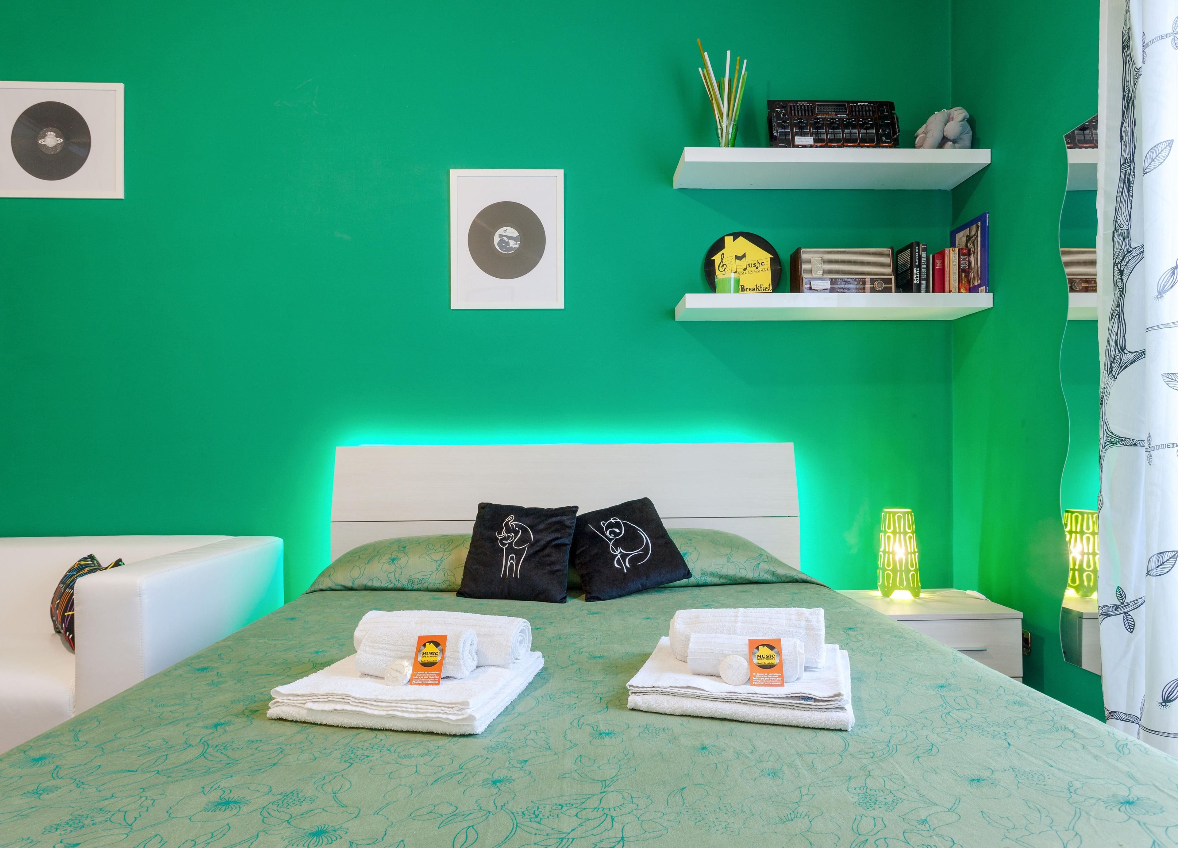 A room with a green wall, a bed with two pillows, a bedside table with a lamp, and a shelf with books and a radio.