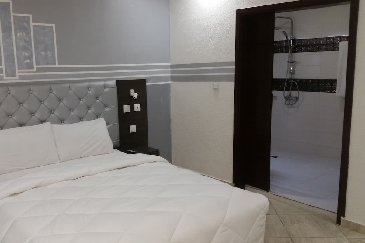 A bedroom with a tufted headboard, a bed with white bedding, a nightstand, a wall-mounted security camera, and a shower area.