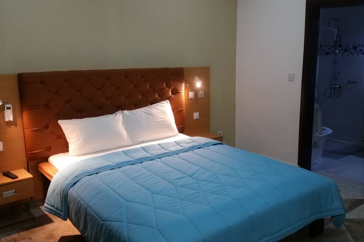 A hotel room with a large bed, a nightstand, a wall-mounted light, and a view of the bathroom.