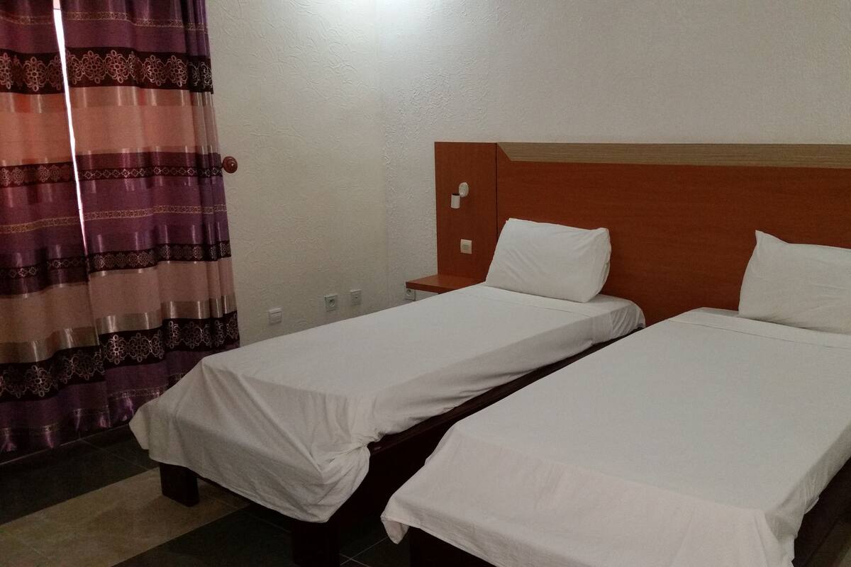 A hotel room with two single beds, a wooden headboard, a nightstand, a curtain, and a wall socket.