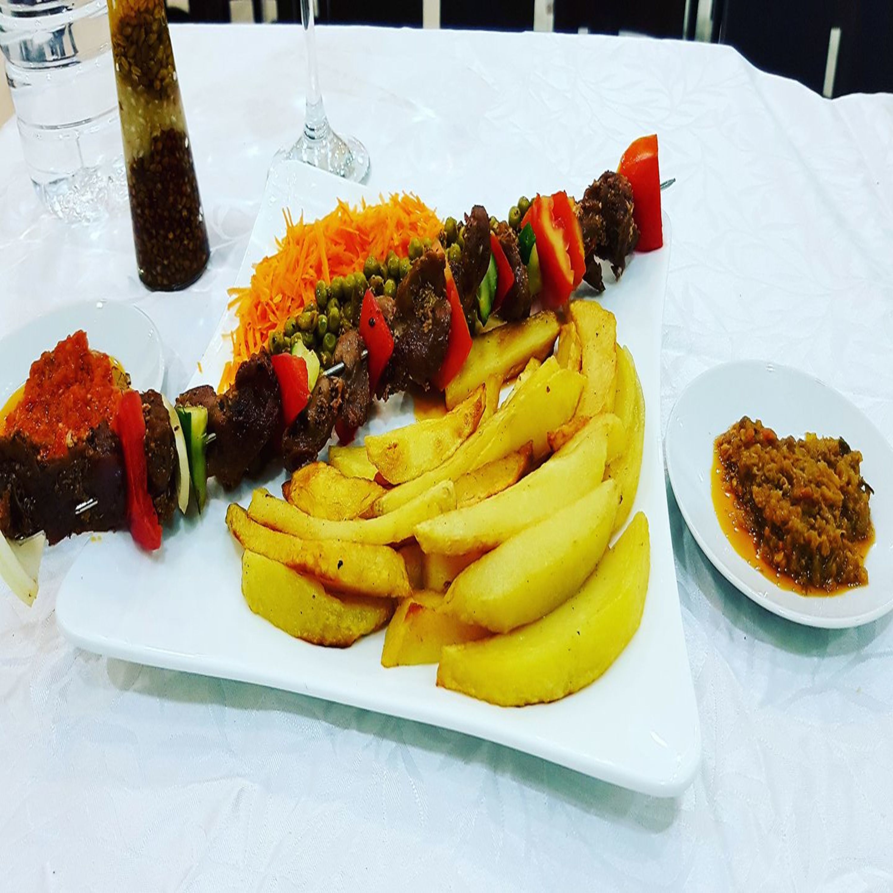 Una mesa blanca con un plato de comida que incluye brochetas, papas fritas y un acompañamiento.