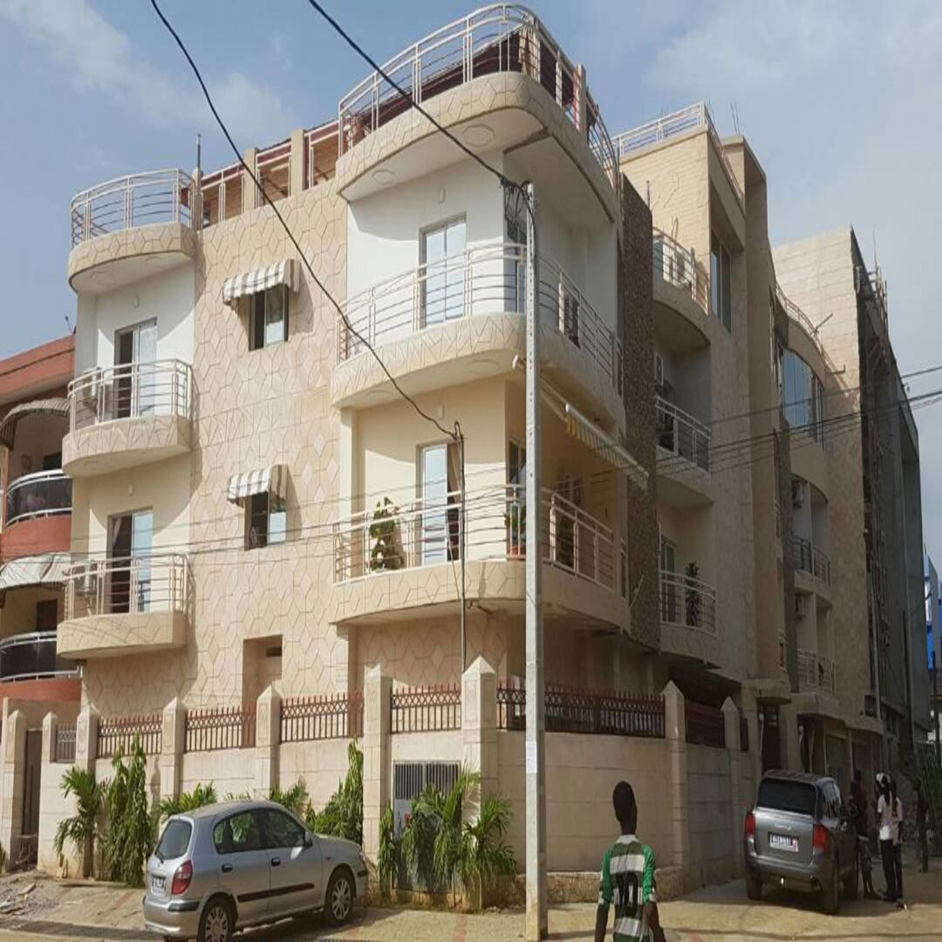 Um edifício residencial com vários andares e varandas, com um carro estacionado na frente e um pedestre em primeiro plano.