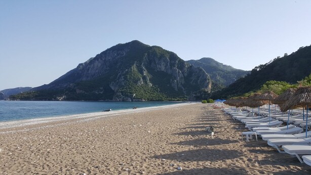 A beach with white lounge chairs and thatched umbrellas, a clear blue sea, and a mountain in the background. - Asya Bungalow (Kemer)