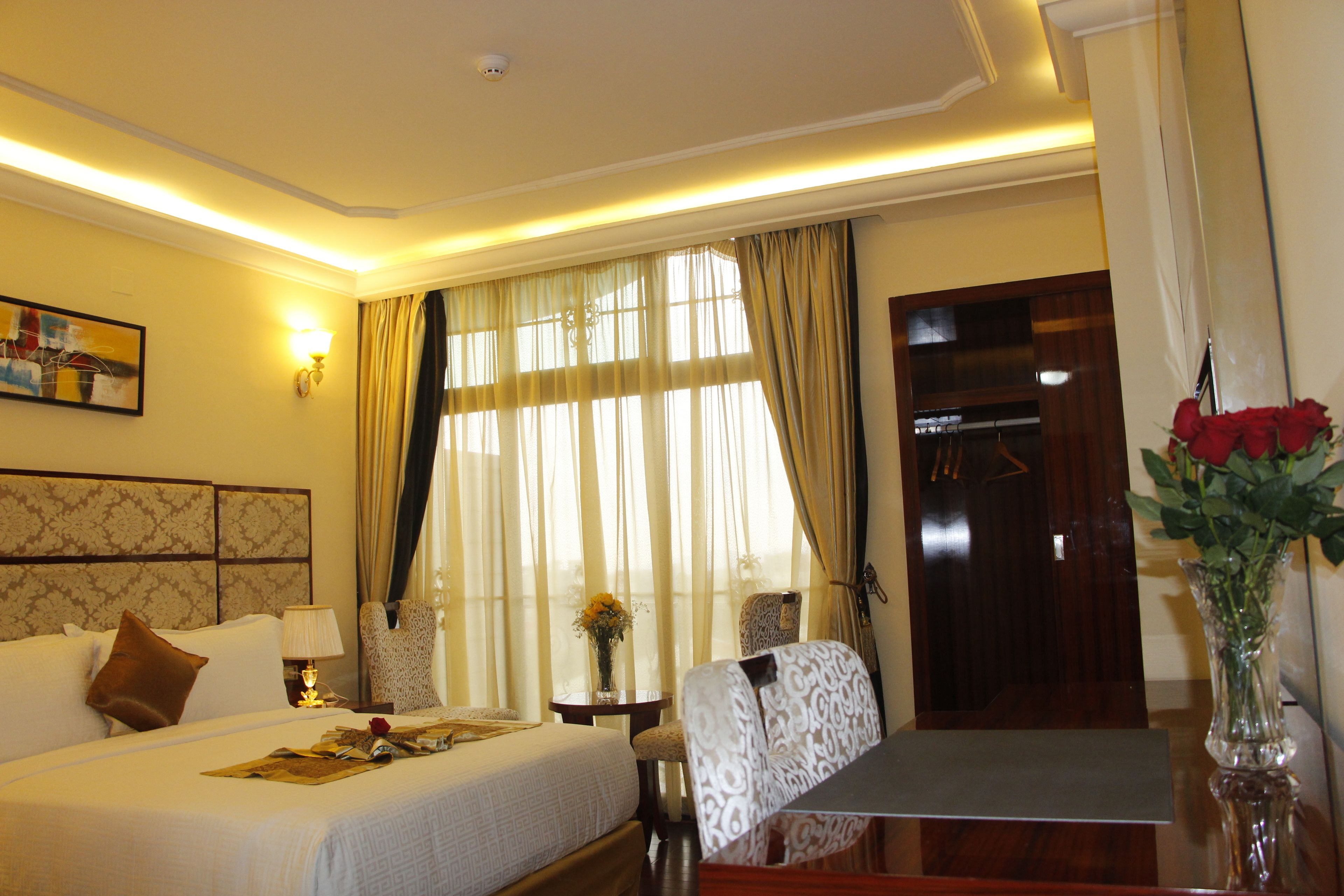 Habitación de hotel con cama, escritorio, silla, un jarrón con flores y ventana con cortinas.