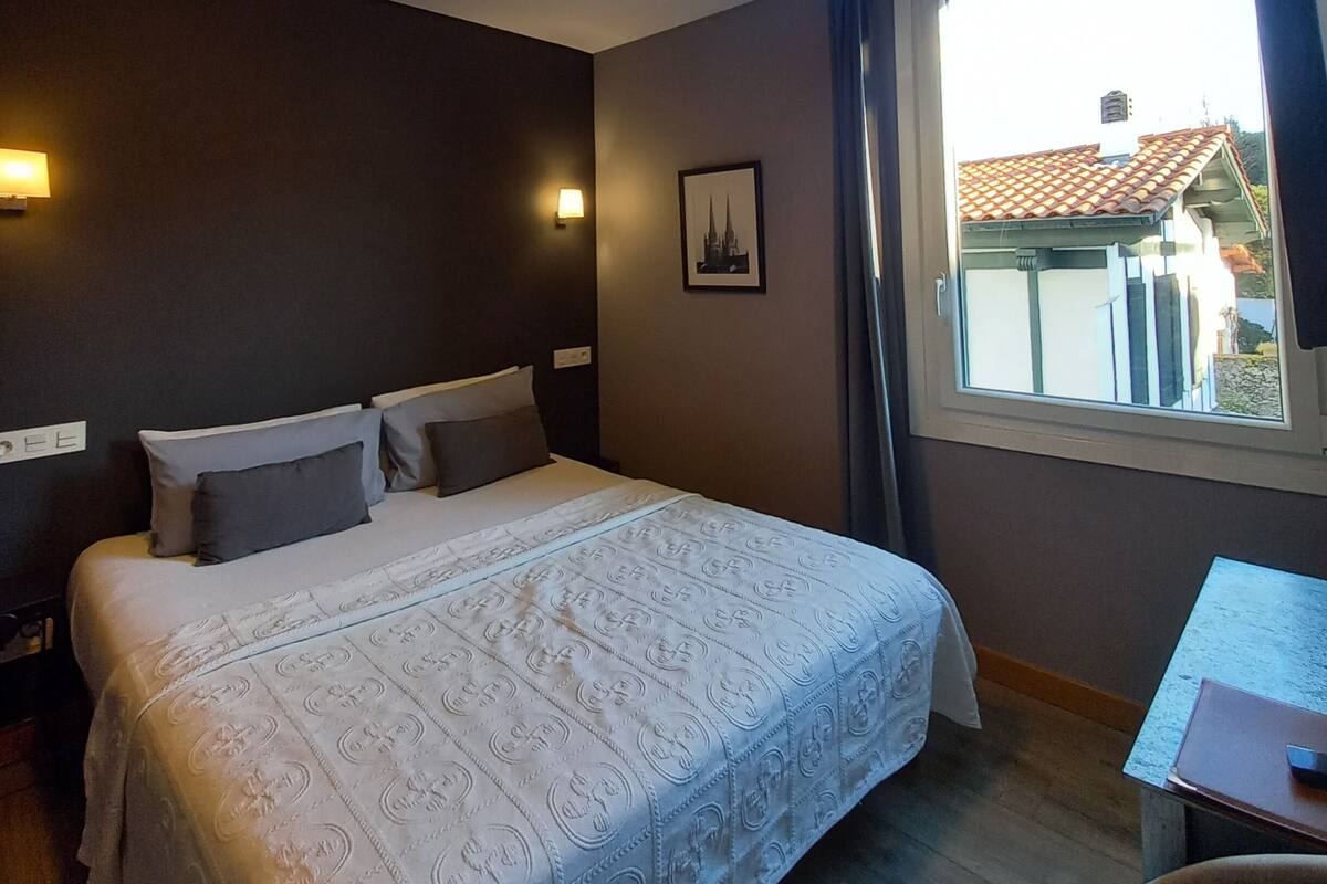 A hotel room with a large bed, bedside lamps, a framed picture on the wall, and a window with a view of a building.