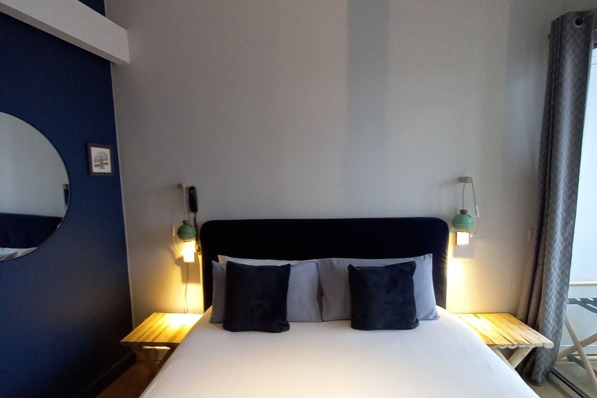A hotel room with a large bed, two bedside tables with lamps, a round mirror on the wall, and a view of the outside.