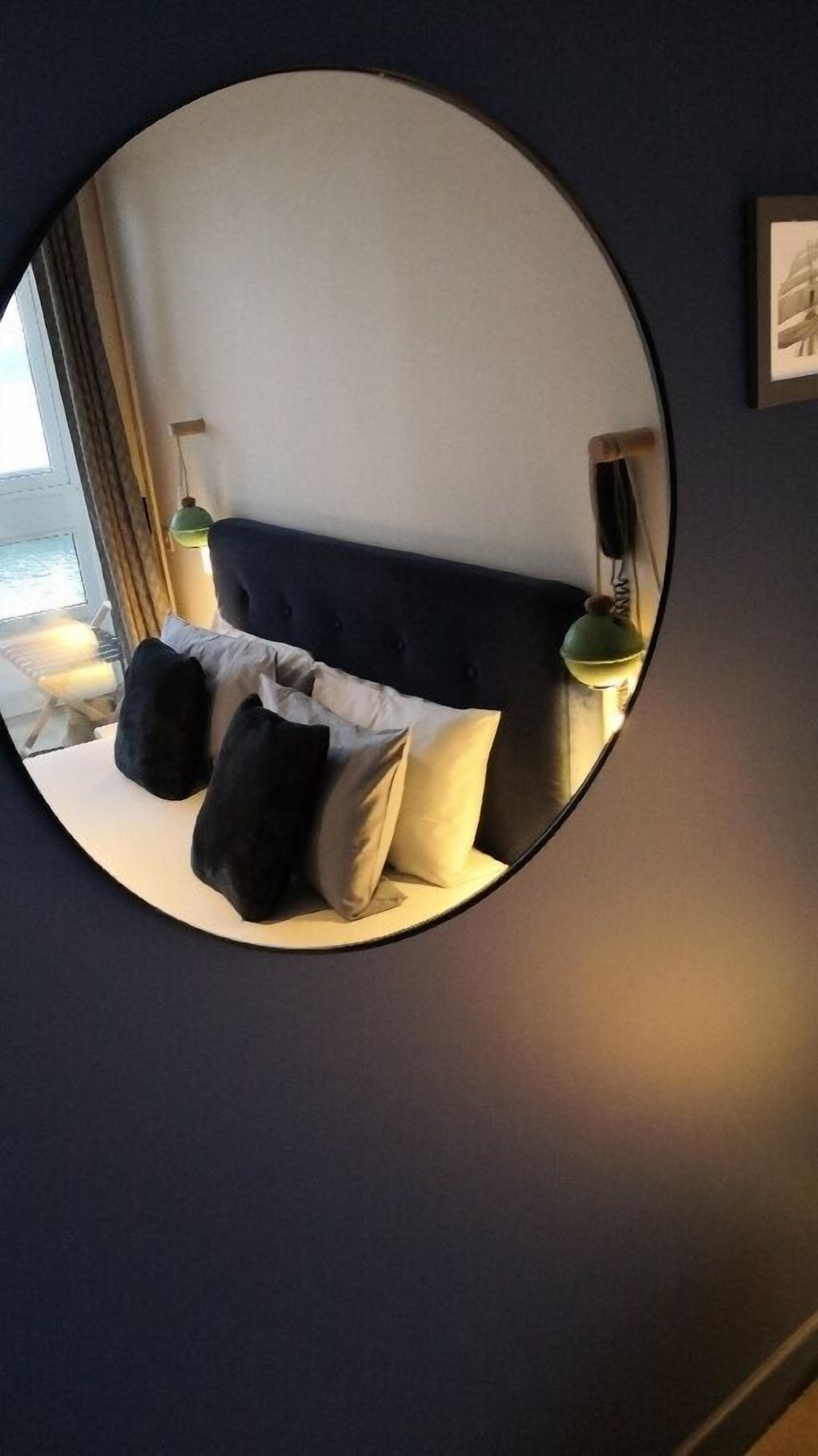 Un espejo circular que refleja una cama con cabecera oscura, varios almohadones y una lámpara de noche.