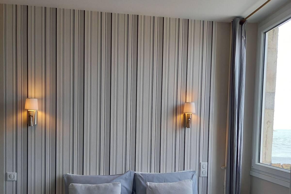A hotel room with a striped wall, a bed with a blue cover, two pillows, two wall-mounted lamps, a nightstand, a window with curtains, and a bench by the window.