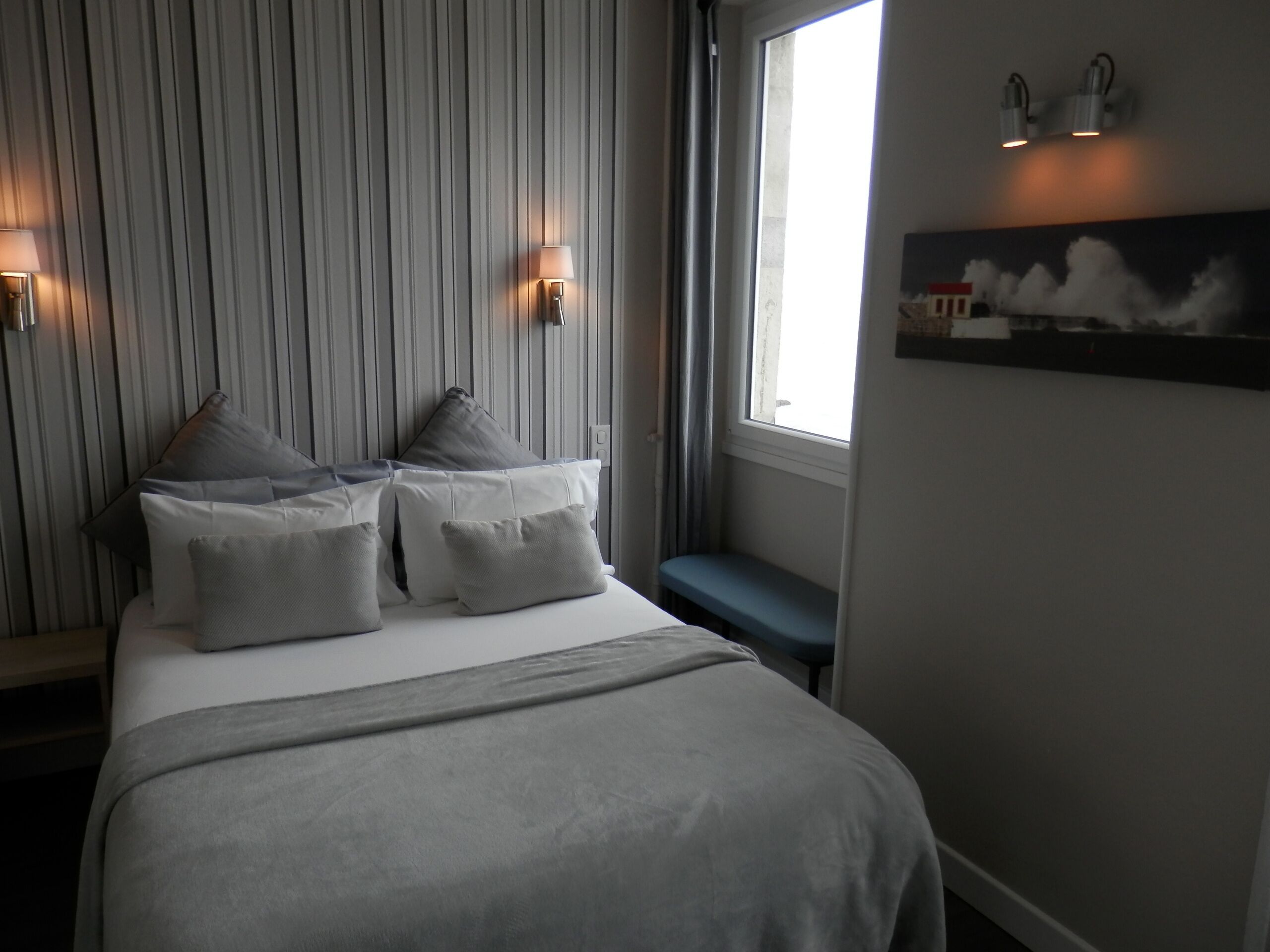 A neatly arranged bedroom with a bed, pillows, a bench, and a wall-mounted lamp.