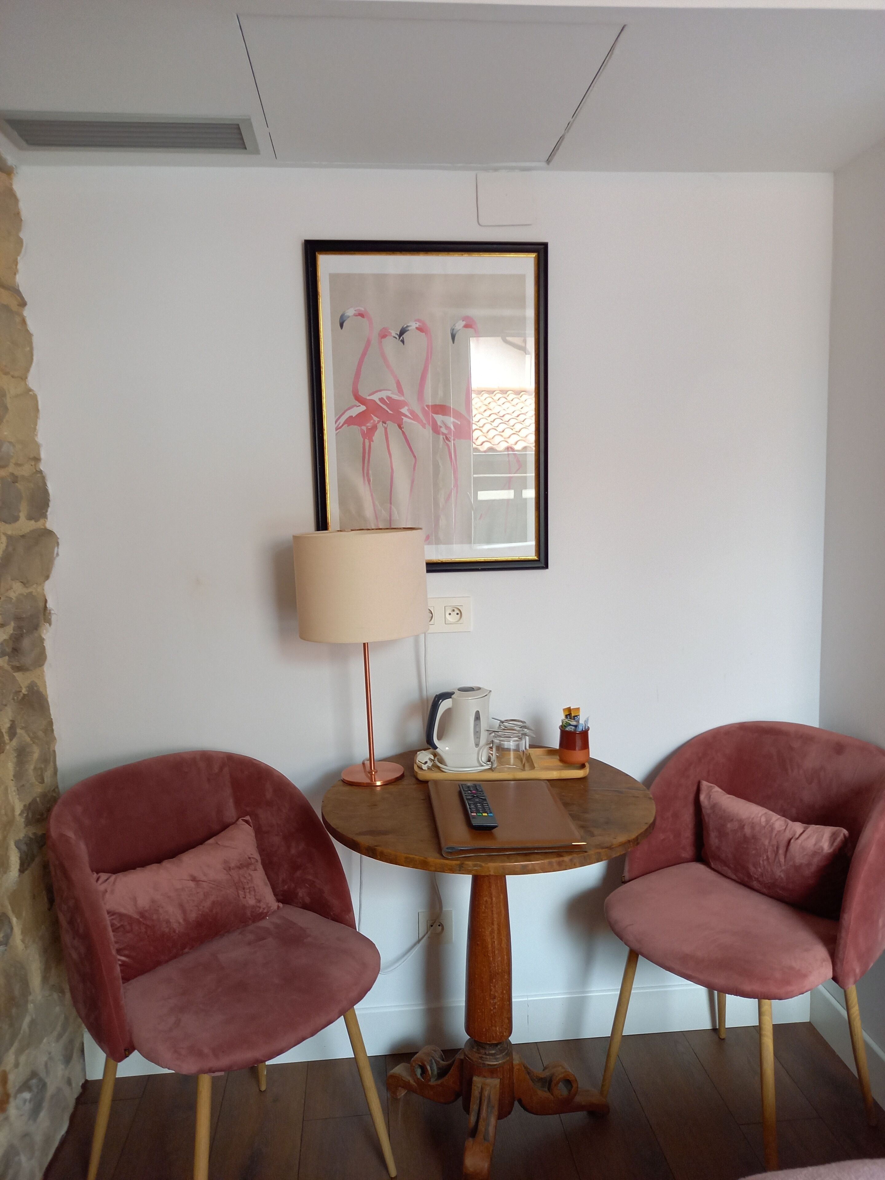 A room with two pink chairs, a wooden table, a lamp, a framed picture, and a kettle.
