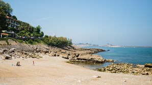 A rocky beach with clear blue water, a few people, and buildings in the background. - La Caravelle (Ciboure)