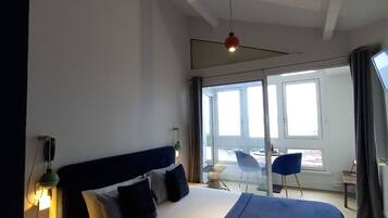 A hotel room with a large bed, a desk, a chair, and a view of the outdoors through a large window.