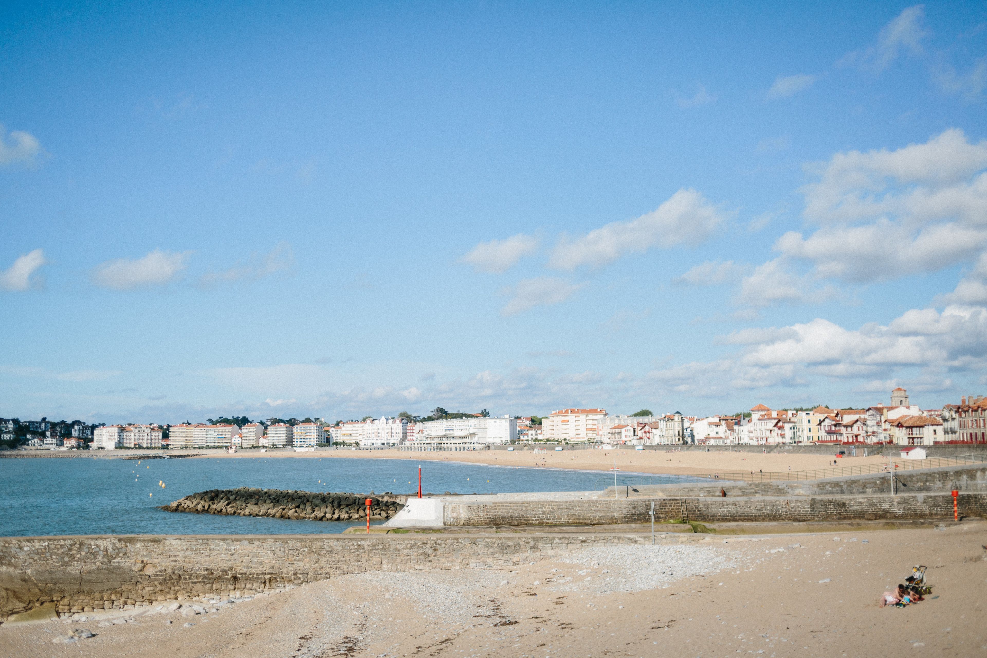 Eine Küstenstadtlandschaft mit Sandstrand, einem Felssteg und Gebäuden mit roten Ziegeldächern.